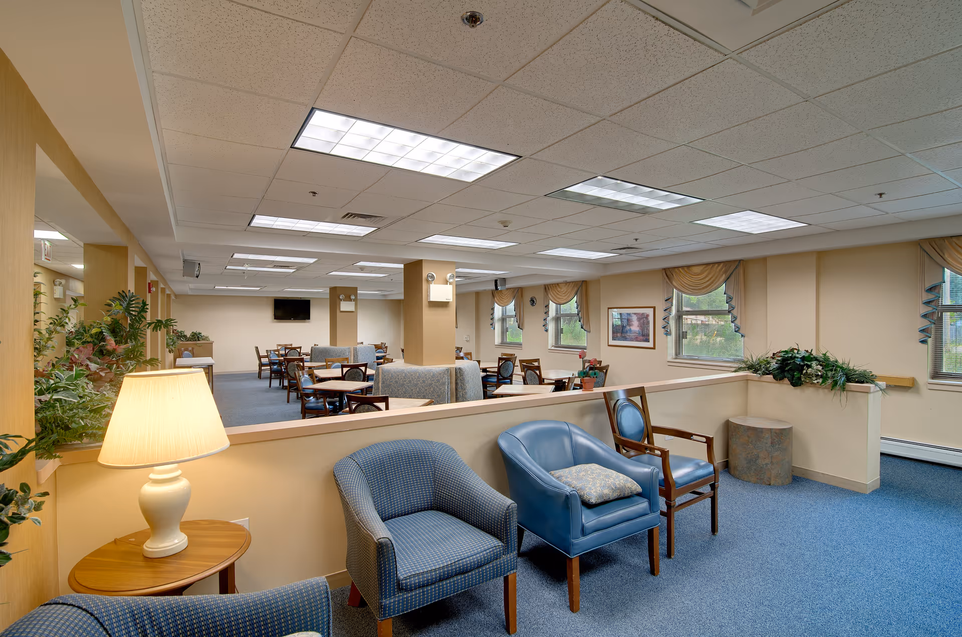 Interior view of a senior living facility common area with multiple seating arrangements including blue and wooden chairs, small tables, and plants. The room has beige walls, blue carpet, several windows with curtains, and fluorescent ceiling lights. A television is mounted on the far wall above a dining area with tables and chairs.