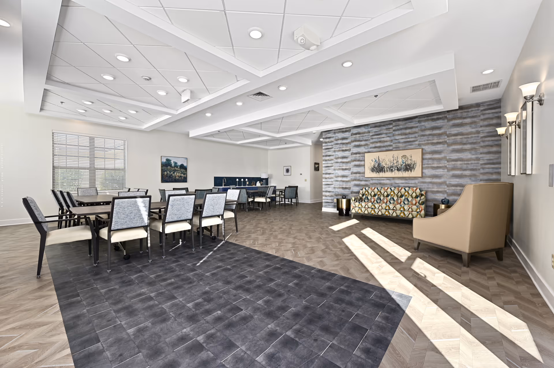 A spacious, well-lit common area with a modern design featuring a dining table with chairs on the left, a patterned sofa and a beige armchair on the right, and a textured accent wall with artwork. The ceiling has recessed lighting and geometric patterns, and sunlight streams in through a window with blinds.