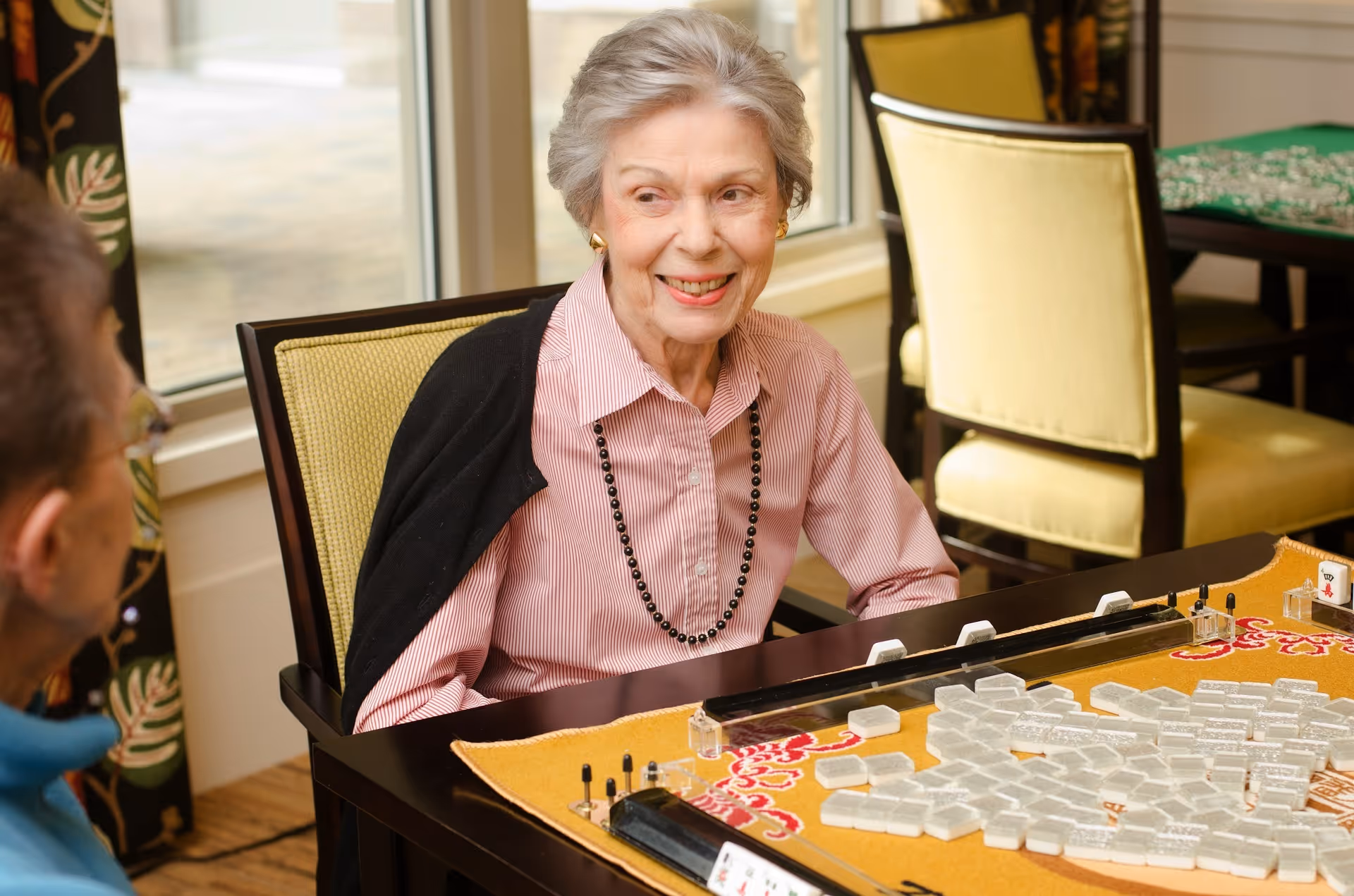 An elderly woman with gray hair, wearing a pink striped shirt and black necklace, sitting at a table playing a game with white tiles, smiling and looking at another person partially visible on the left. The room has large windows and yellow cushioned chairs.