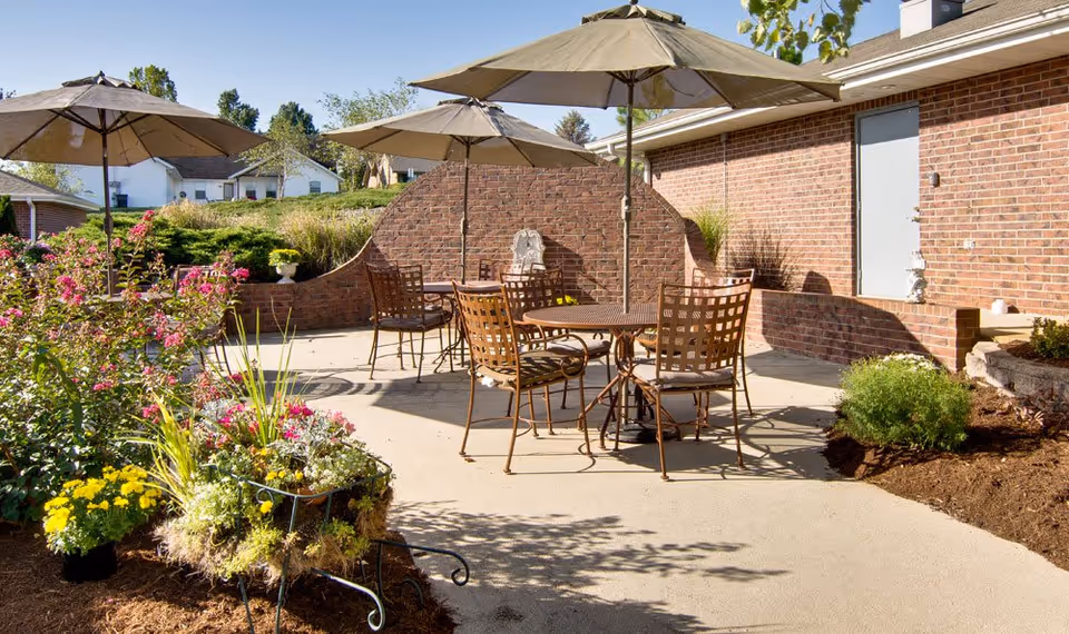 Outdoor patio area with metal tables and chairs under large beige umbrellas, surrounded by flower beds with colorful plants and a brick wall in the background.