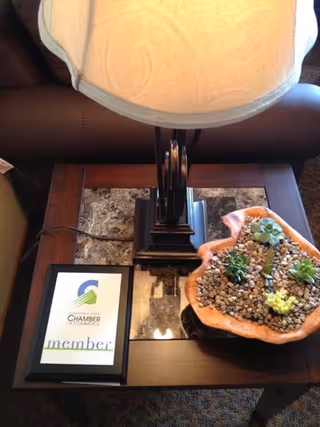 A close-up view of a wooden side table with a marble inset, featuring a table lamp with a beige shade, a framed Chamber of Commerce member certificate, and a decorative bowl filled with small pebbles and succulent plants.