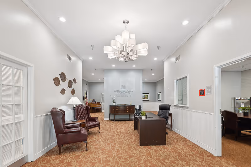 Interior view of a reception or lobby area at Cameron Hall of Canton featuring two leather armchairs on the left, a dark wood desk with an office chair on the right, a decorative chandelier hanging from the ceiling, and a cabinet with plants and decor against the back wall with the facility name displayed.