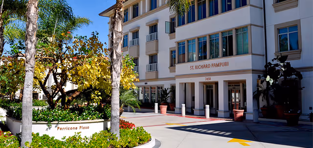 Exterior view of the St. Richard Pampuri senior living facility showing the building entrance with the name displayed above it. The foreground features a landscaped area with palm trees, green shrubs, and a sign that reads Perricone Plaza under a clear blue sky.