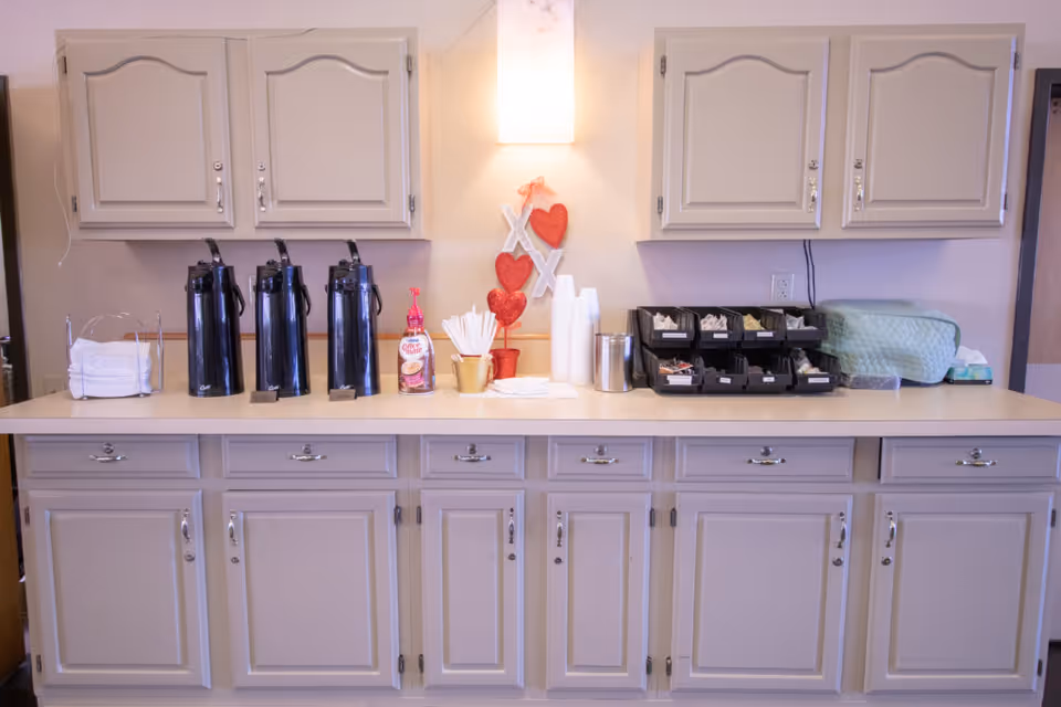 Countertop coffee station with three thermal dispensers, disposable cups, sweeteners and cabinets in a communal kitchenette.