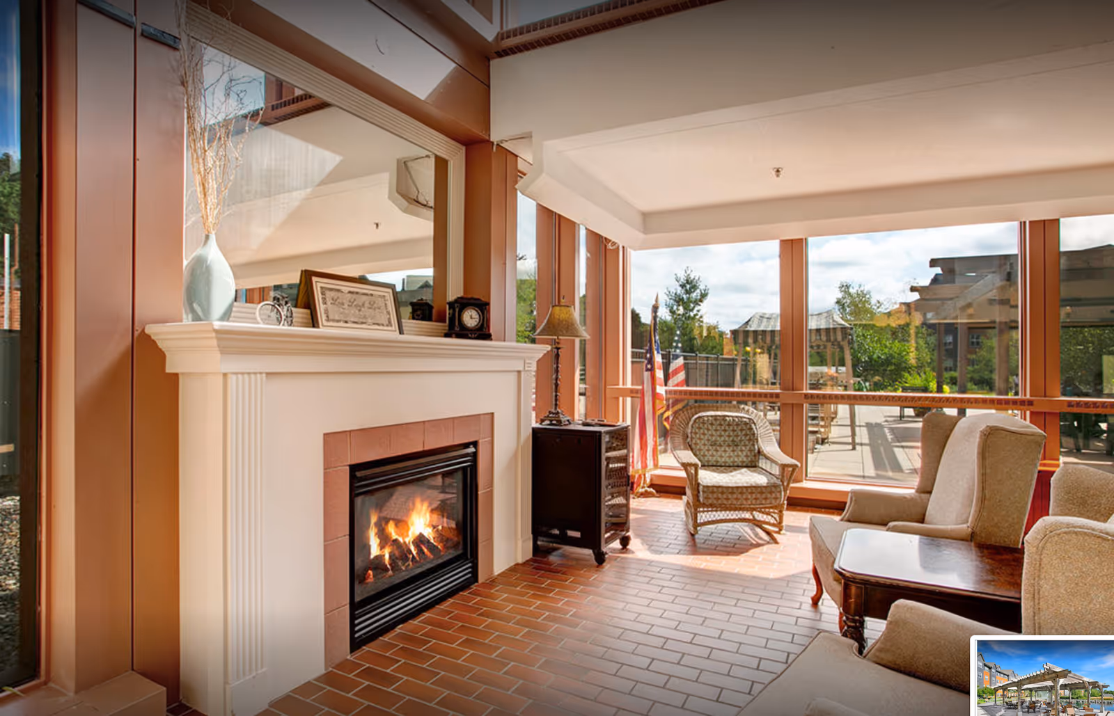 A cozy sitting area with a lit fireplace, a large mirror above the mantel, a wicker chair, two upholstered armchairs, a wooden side table, and large windows showing an outdoor patio with pergolas and greenery.