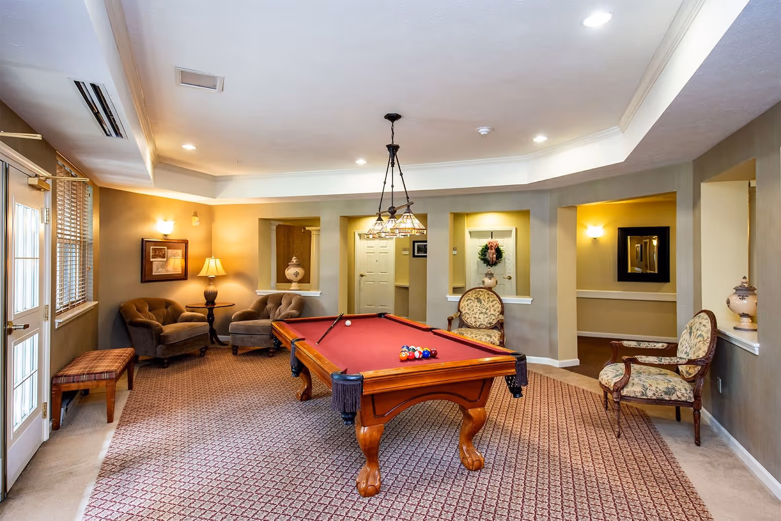 A cozy game room with a red felt pool table in the center, surrounded by upholstered chairs and armchairs. The room has beige walls, carpeted floor with a patterned design, and soft lighting from wall sconces and a hanging light fixture above the pool table. There are decorative vases on shelves built into the walls and a framed picture on one wall. A door with glass panels and window blinds is visible on the left side.