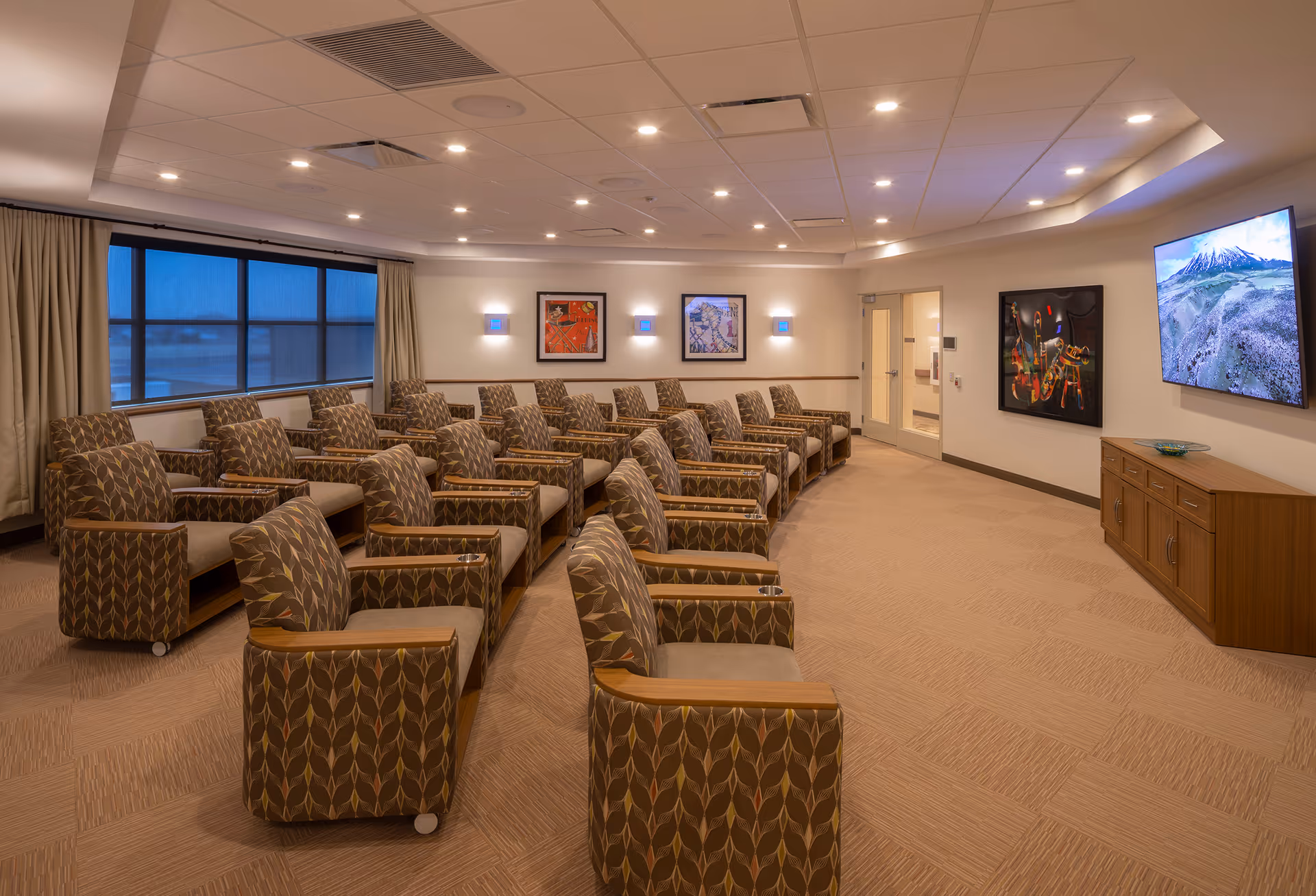 A spacious senior living media room with rows of patterned armchairs facing a wall-mounted TV and artwork.