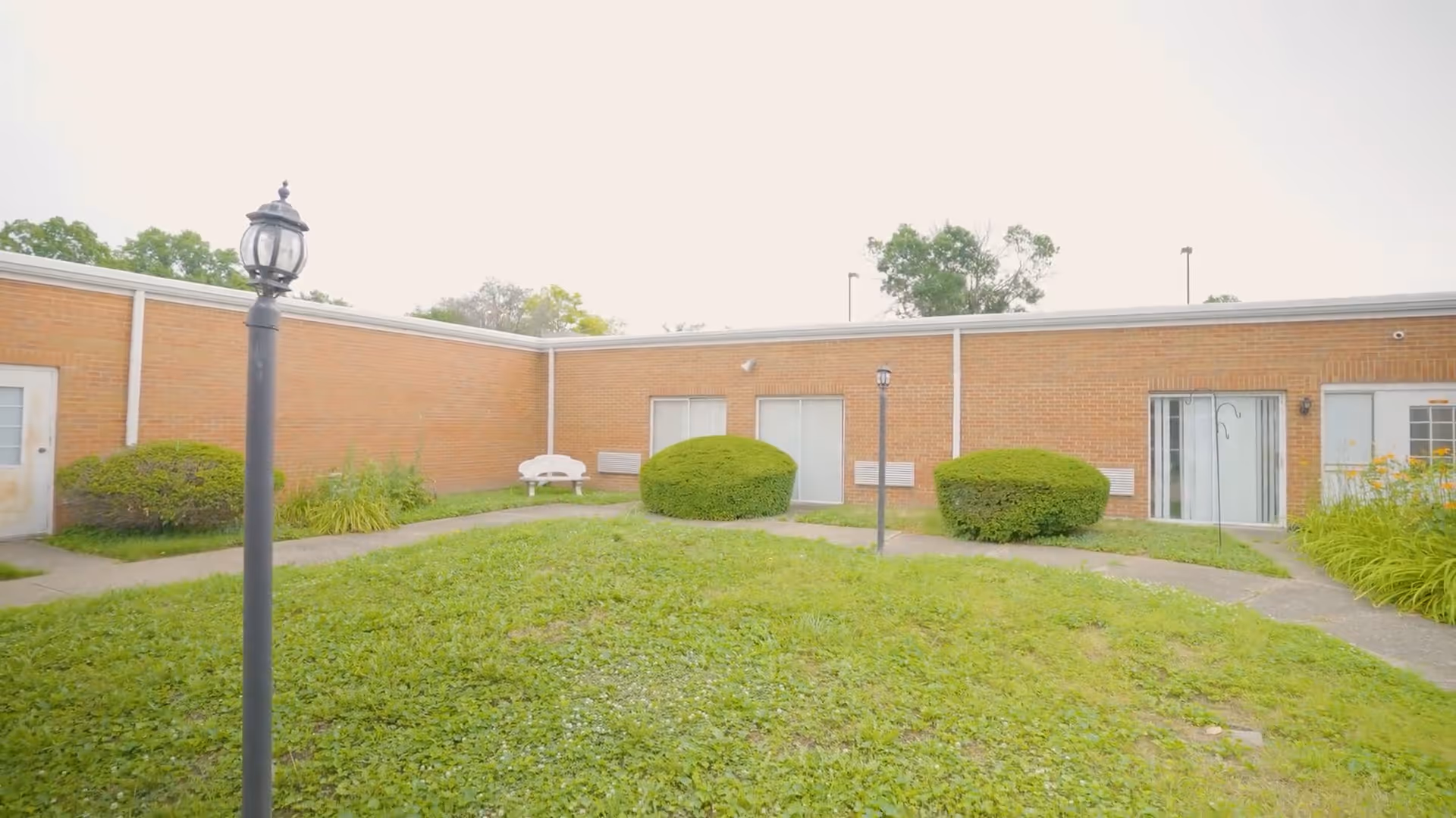 Brick single-story courtyard with grass, trimmed shrubs, lamp posts, a white bench and sliding glass doors.