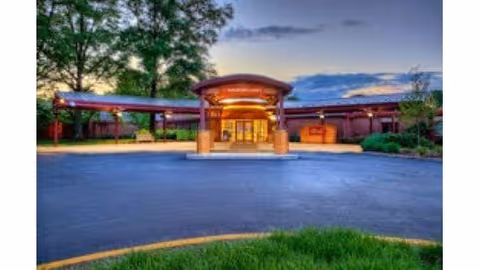 Entrance of a building at dusk with a covered driveway, surrounded by trees and greenery under a partly cloudy sky.