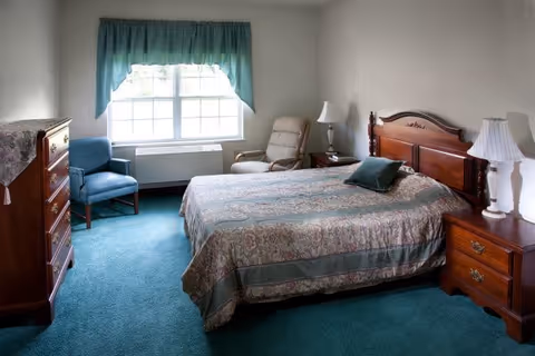A tidy bedroom with a bed, wooden headboard, matching nightstands and dresser, two chairs, and a window with teal curtains.