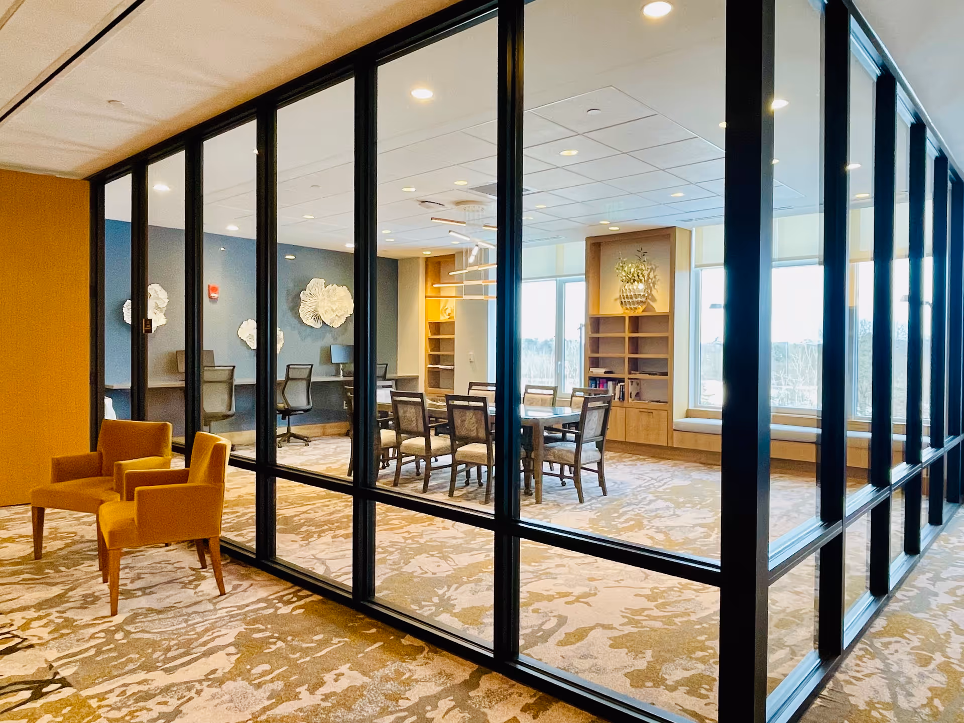 A modern senior living facility interior featuring a glass-walled meeting or dining room with a table and chairs inside. Outside the glass walls, there are two mustard-colored armchairs on a patterned carpet. The room inside has large windows with natural light, built-in shelves, and decorative wall art.