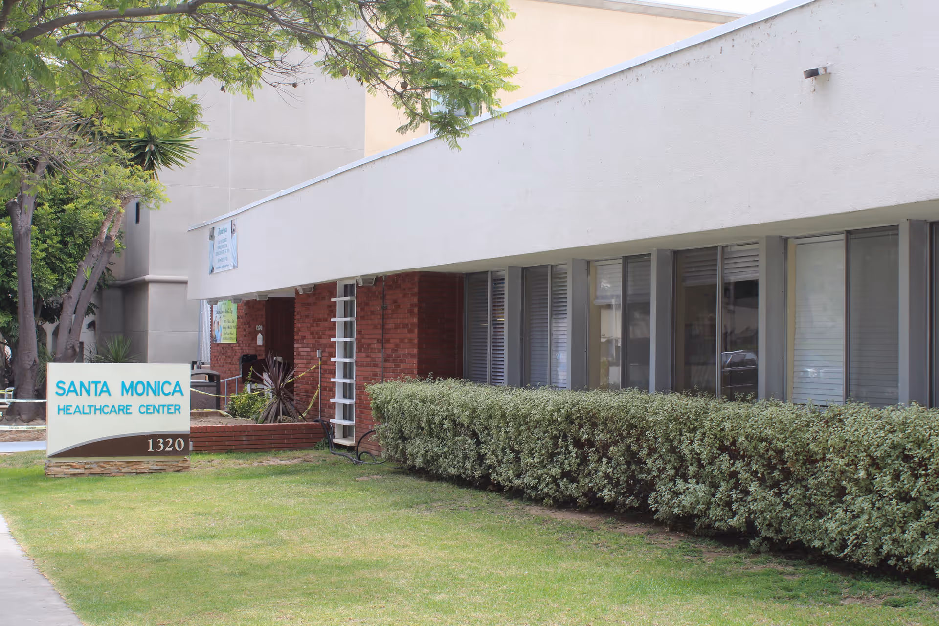 Exterior view of Santa Monica Healthcare Center building with a sign displaying the facility name and address 1320. The building has large windows with bushes in front and a tree on the left side.