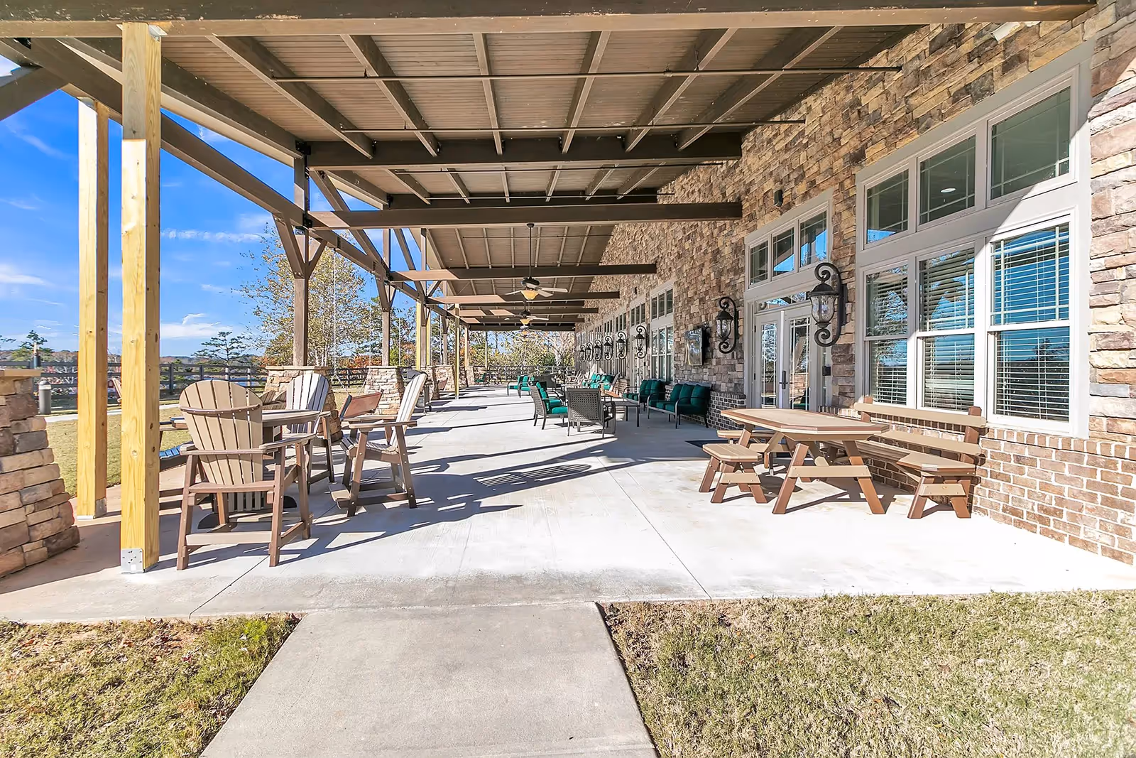 A spacious covered outdoor patio area with wooden beams and ceiling fans. The patio features multiple seating arrangements including wooden chairs with small tables, green cushioned sofas, and a wooden picnic table with benches. The building wall is made of stone and brick with several windows and decorative wall-mounted lanterns. The patio overlooks a grassy area under a clear blue sky.
