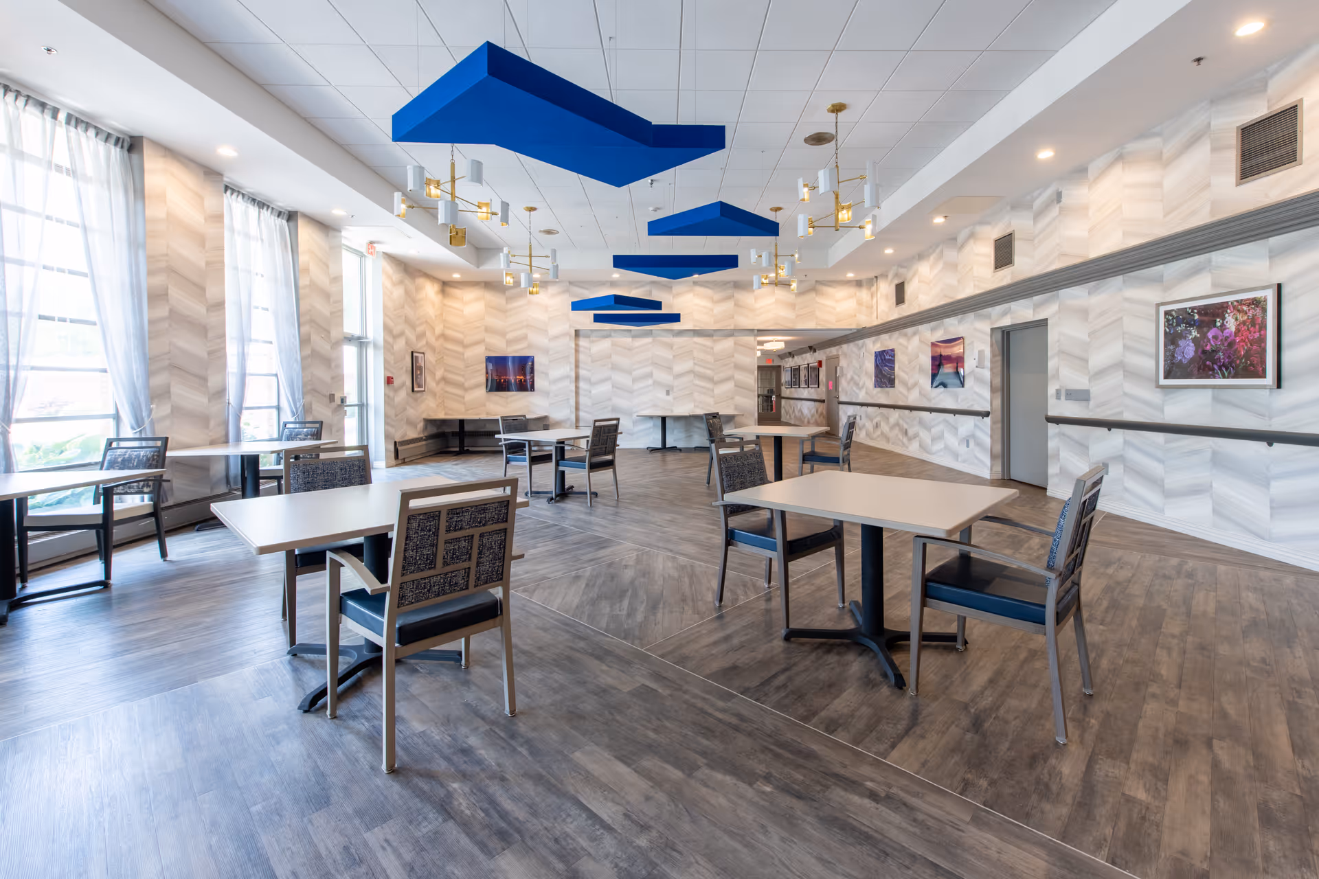 Bright communal dining/activity room with tables and chairs, large windows, patterned walls, and blue ceiling panels.