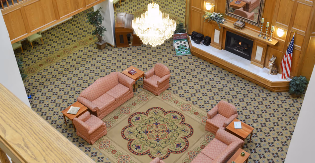 Overhead view of a senior living facility lounge area with patterned carpet, a large decorative rug, multiple pink upholstered sofas and armchairs arranged around wooden side tables, a large chandelier, a fireplace with a mirror above it, an American flag, and some plants.