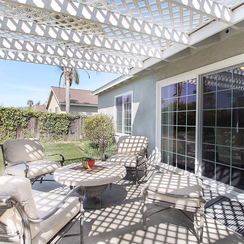 Sunlit backyard patio with a white lattice pergola casting grid shadows over cushioned chairs and a round table next to sliding glass doors.