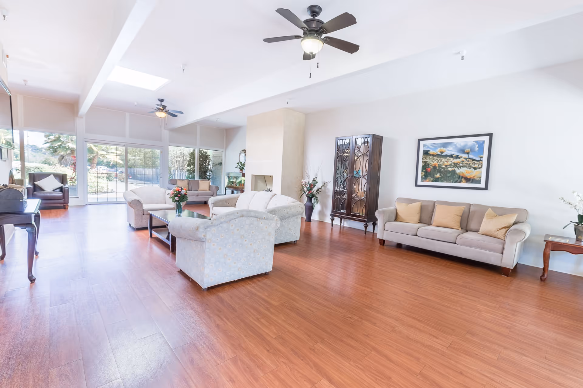A spacious and bright living room with wooden flooring, multiple sofas and armchairs arranged around a coffee table with a vase of flowers, a ceiling fan with light, large windows and sliding glass doors letting in natural light, a decorative cabinet, and a framed floral painting on the wall.