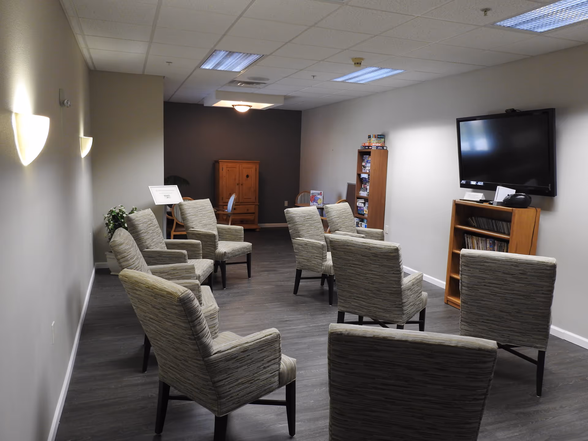 A common room with eight upholstered chairs arranged in a semi-circle facing a wall-mounted flat screen TV. The room has wood flooring, neutral-colored walls, and ceiling lights. In the background, there is a wooden cabinet, a bookshelf filled with board games, and a small table with chairs.