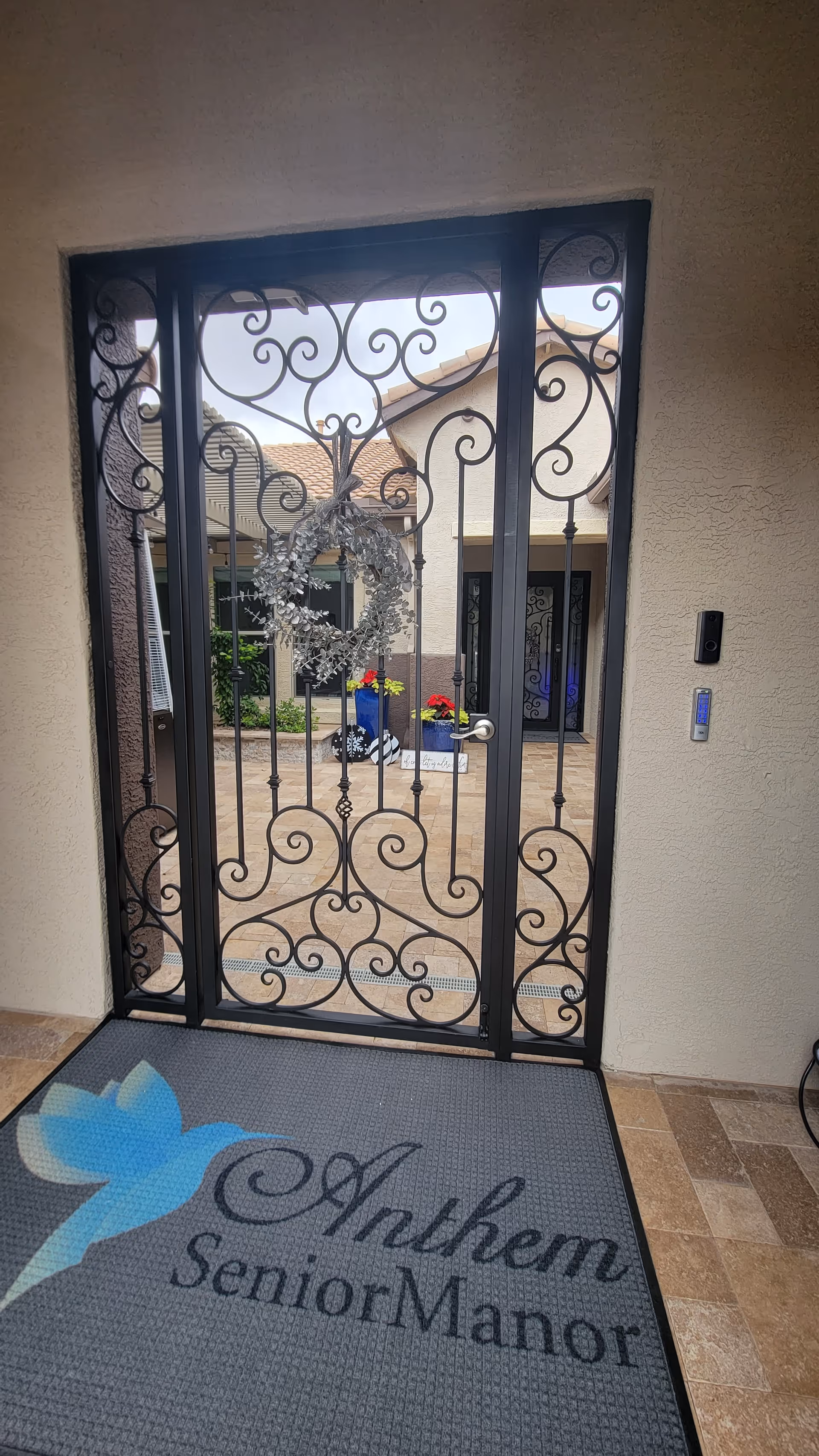 View through a decorative wrought iron gate with a silver wreath hanging on it, looking out onto a courtyard with potted plants and a building entrance. A doormat with a blue hummingbird and the text 'Anthem Senior Manor' is visible in the foreground.