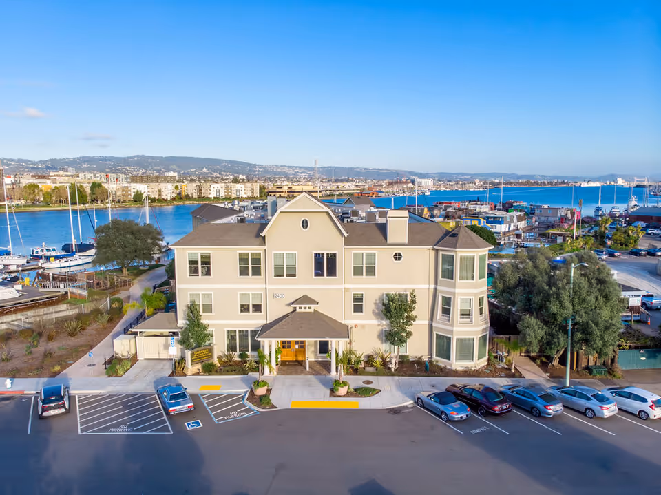 Exterior view of a three-story beige building with multiple windows and a central entrance, located near a waterfront with boats docked. There is a parking lot with several cars in front of the building and a clear blue sky above.