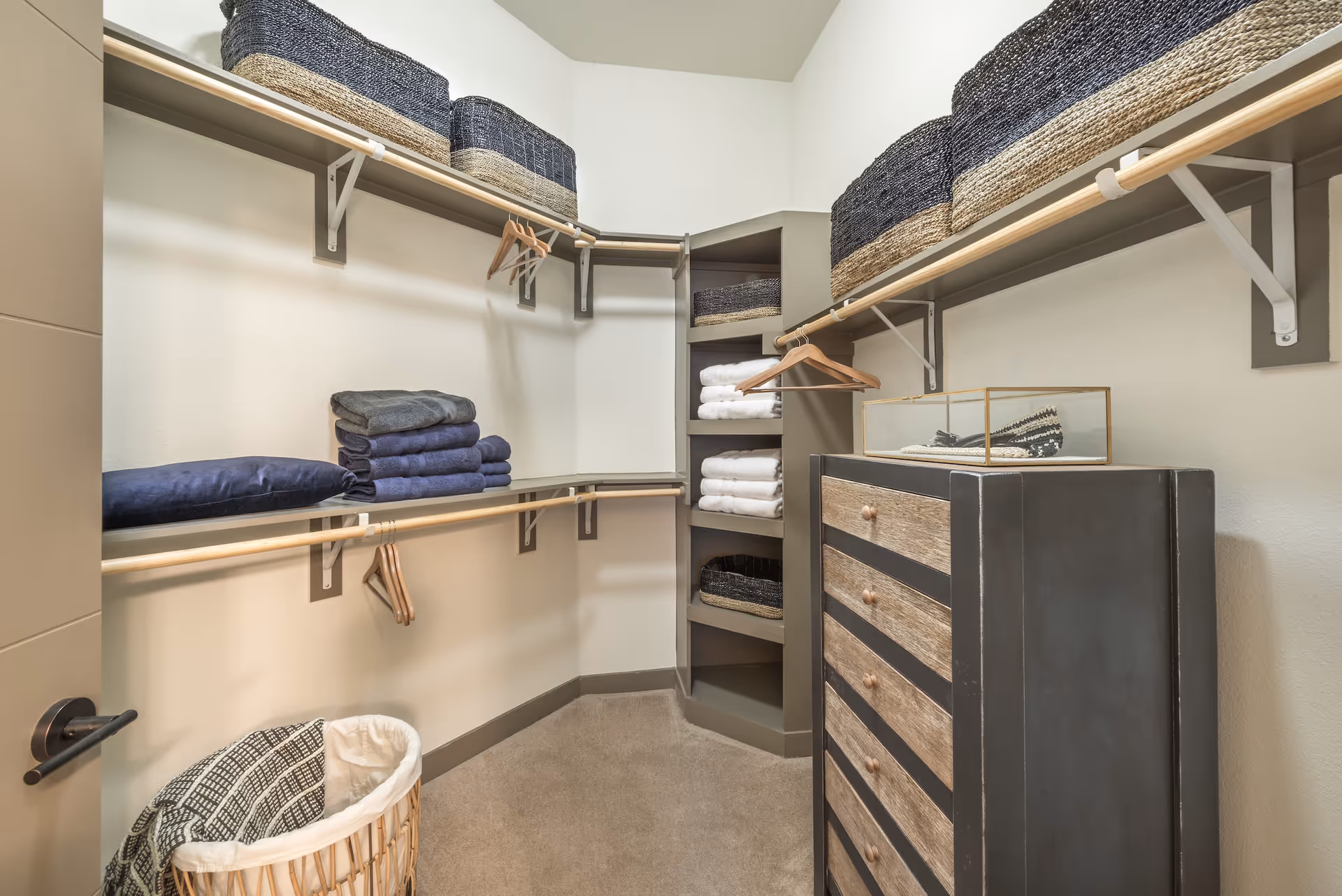 Well-organized walk-in closet with shelving, hanging rods, woven baskets, folded towels and a chest of drawers.