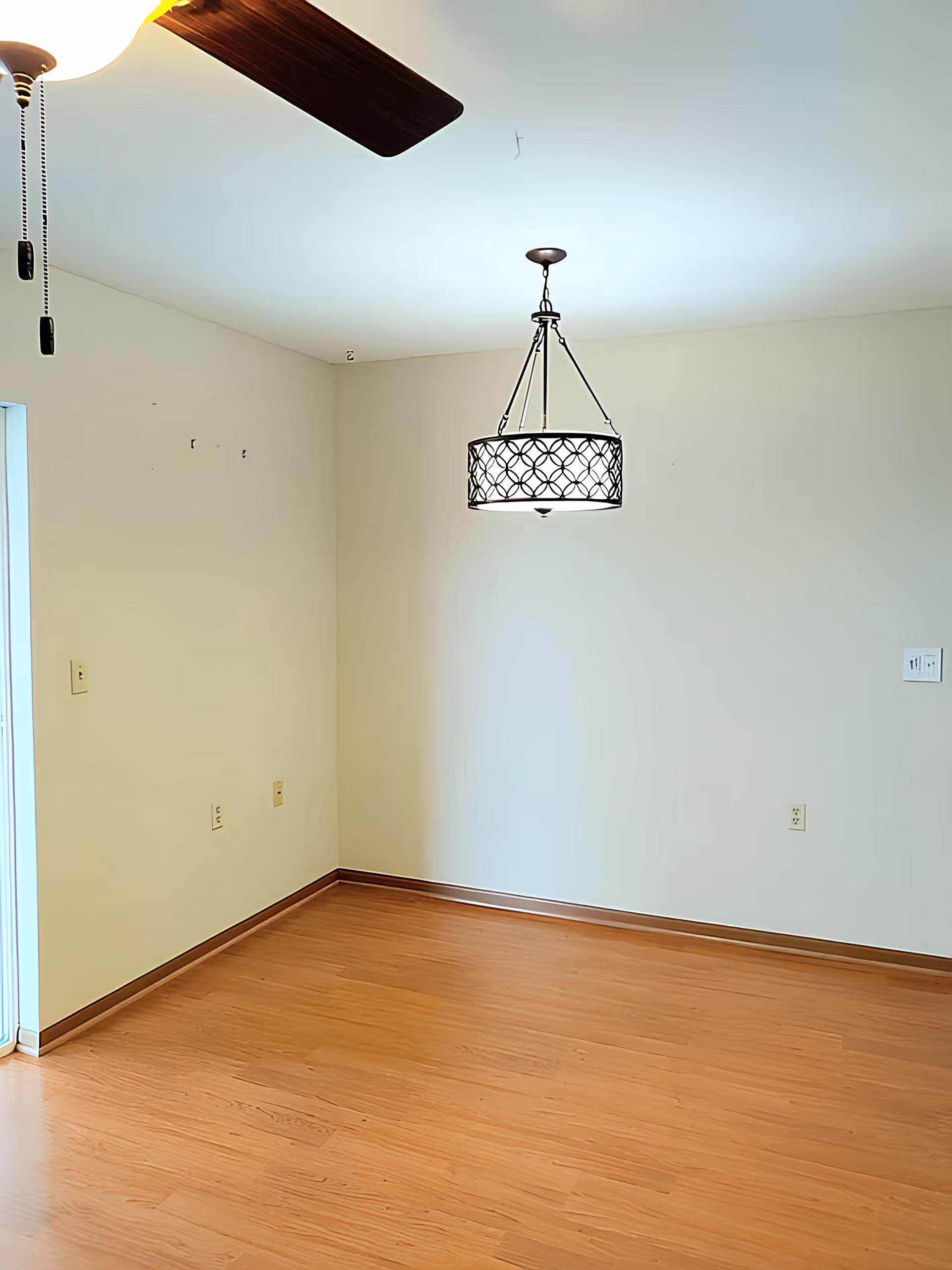 Empty room with wood floors, cream-colored walls, a hanging decorative light fixture, and a ceiling fan blade visible.