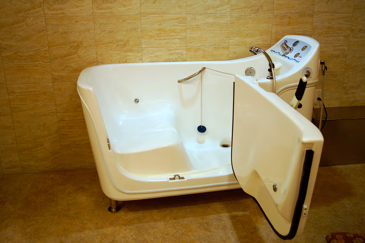 A white walk-in bathtub with an open door, designed for easy access, placed against a beige tiled wall and floor. The bathtub has built-in controls and a handheld showerhead attached.