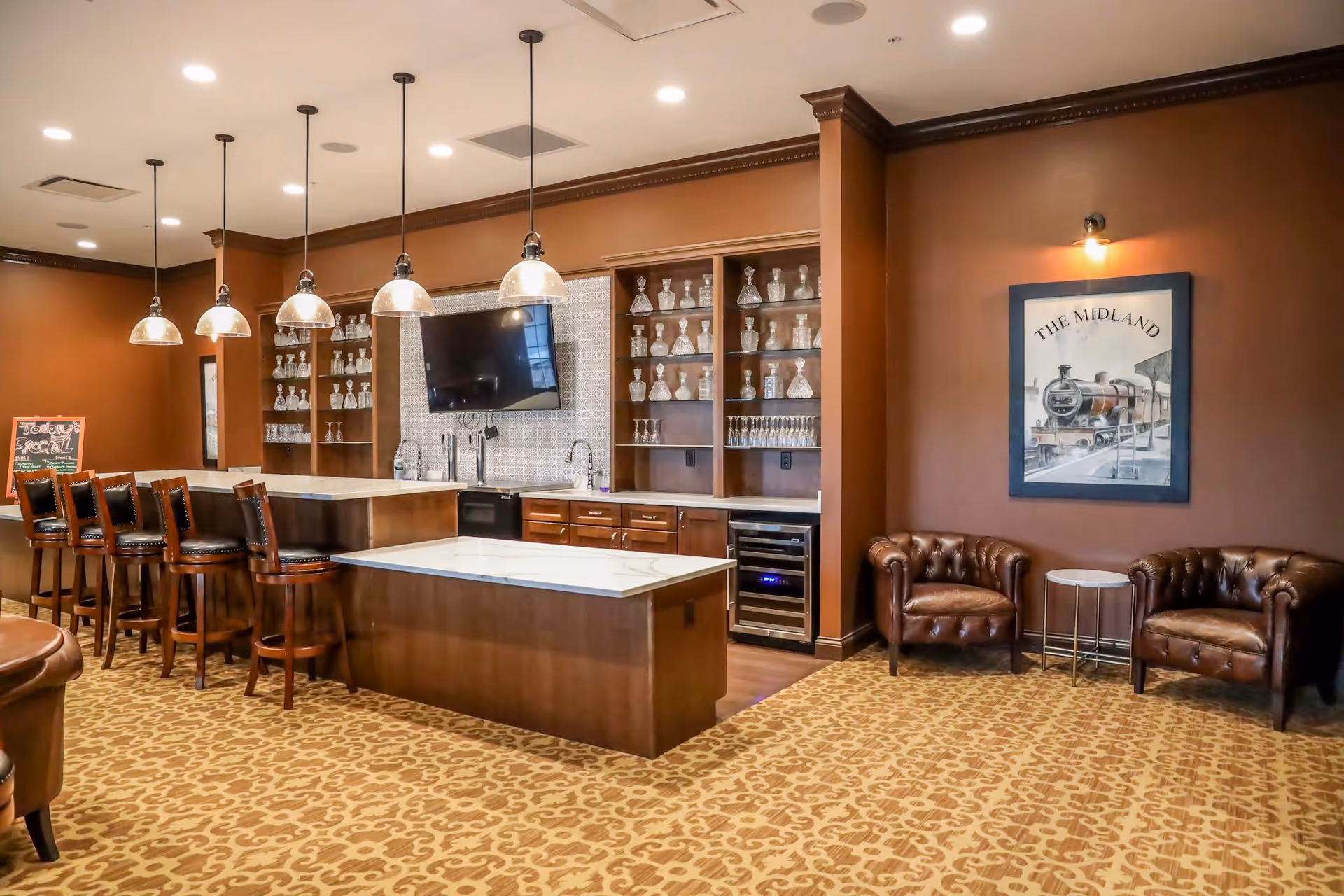A cozy lounge area with a bar counter featuring five wooden bar stools, pendant lights hanging above, and shelves filled with glassware. There is a flat-screen TV mounted on the wall behind the bar. To the right, two brown leather armchairs flank a small round table beneath a framed picture of a train labeled 'The Midland.' The room has warm brown walls and patterned carpet flooring.