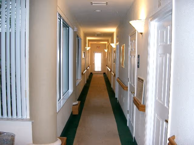 A long, narrow hallway in a senior living facility with beige walls, green carpet borders, and a central beige carpet runner. The hallway has white doors on the right side, windows with vertical blinds on the left, wall-mounted lights, and handrails along both sides. At the end of the hallway, there is a door with a window letting in natural light.