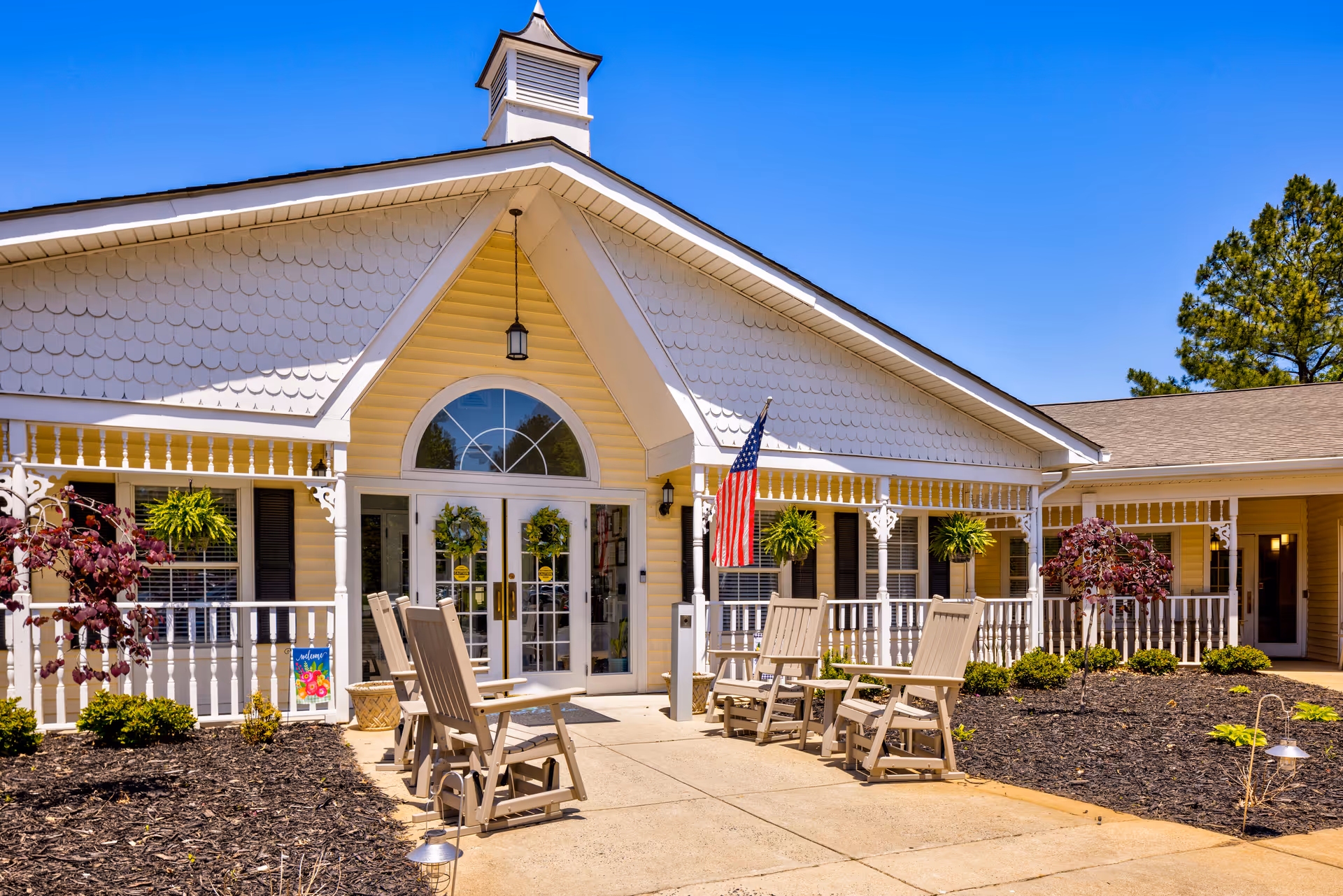 Exterior view of TerraBella Harrisburg facility showing a single-story building with a peaked roof, white railings, hanging plants, and an American flag. There are several wooden rocking chairs arranged on a concrete patio in front of the entrance under a clear blue sky.