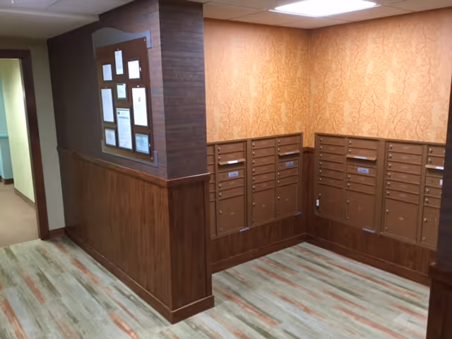 Interior corner of a senior living facility hallway with multiple brown mailboxes mounted on the walls, a bulletin board with notices, wood paneling on the lower half of the walls, patterned wallpaper above, and a light-colored wood floor.