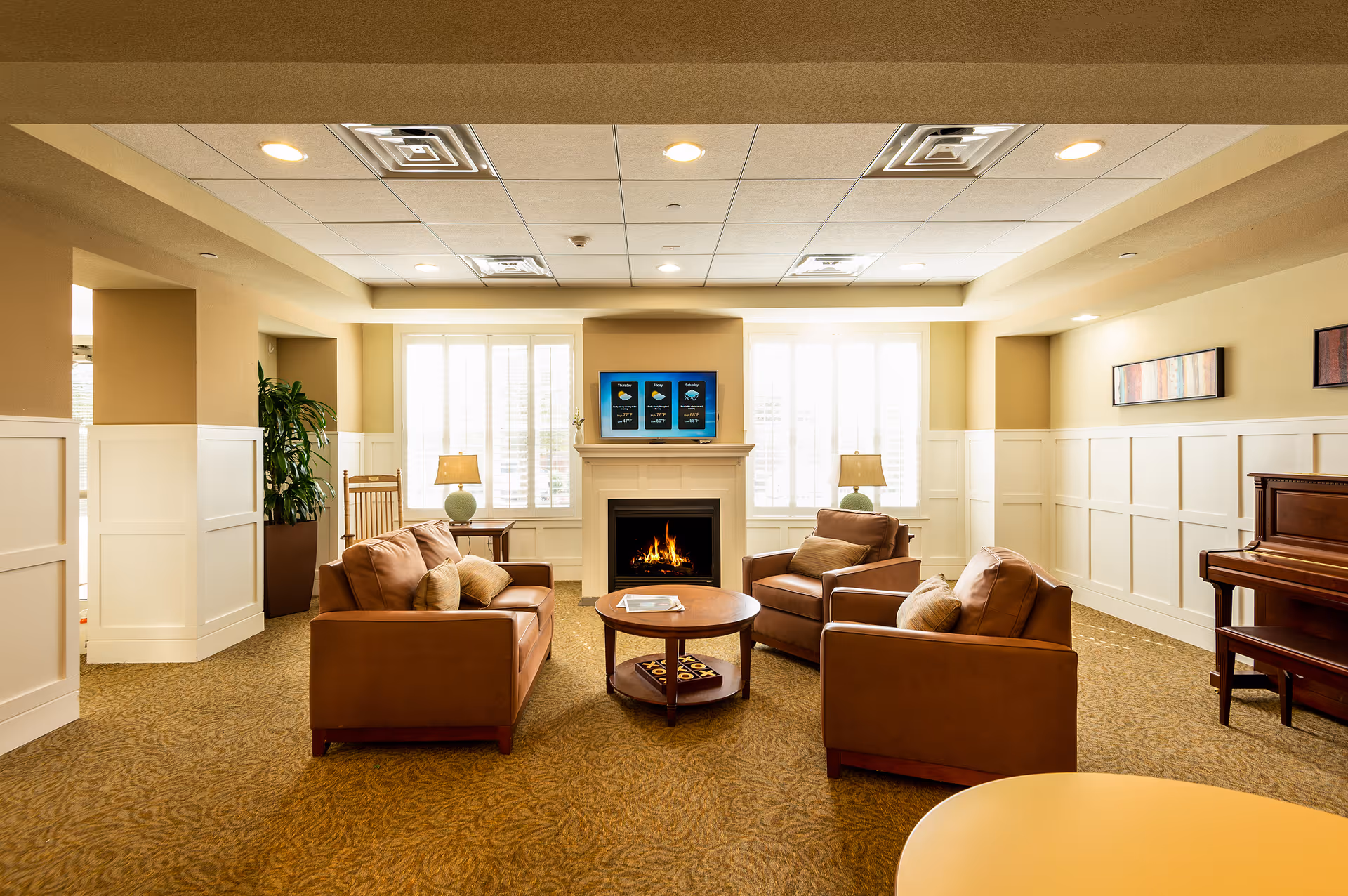 A cozy living room area with three brown leather chairs and a matching loveseat arranged around a round wooden coffee table. There is a fireplace with a TV mounted above it displaying a weather forecast. Two table lamps with green bases sit on side tables next to the chairs. The room has beige walls with white wainscoting, a patterned carpet, and a piano on the right side. Large windows with white shutters allow natural light to fill the space.