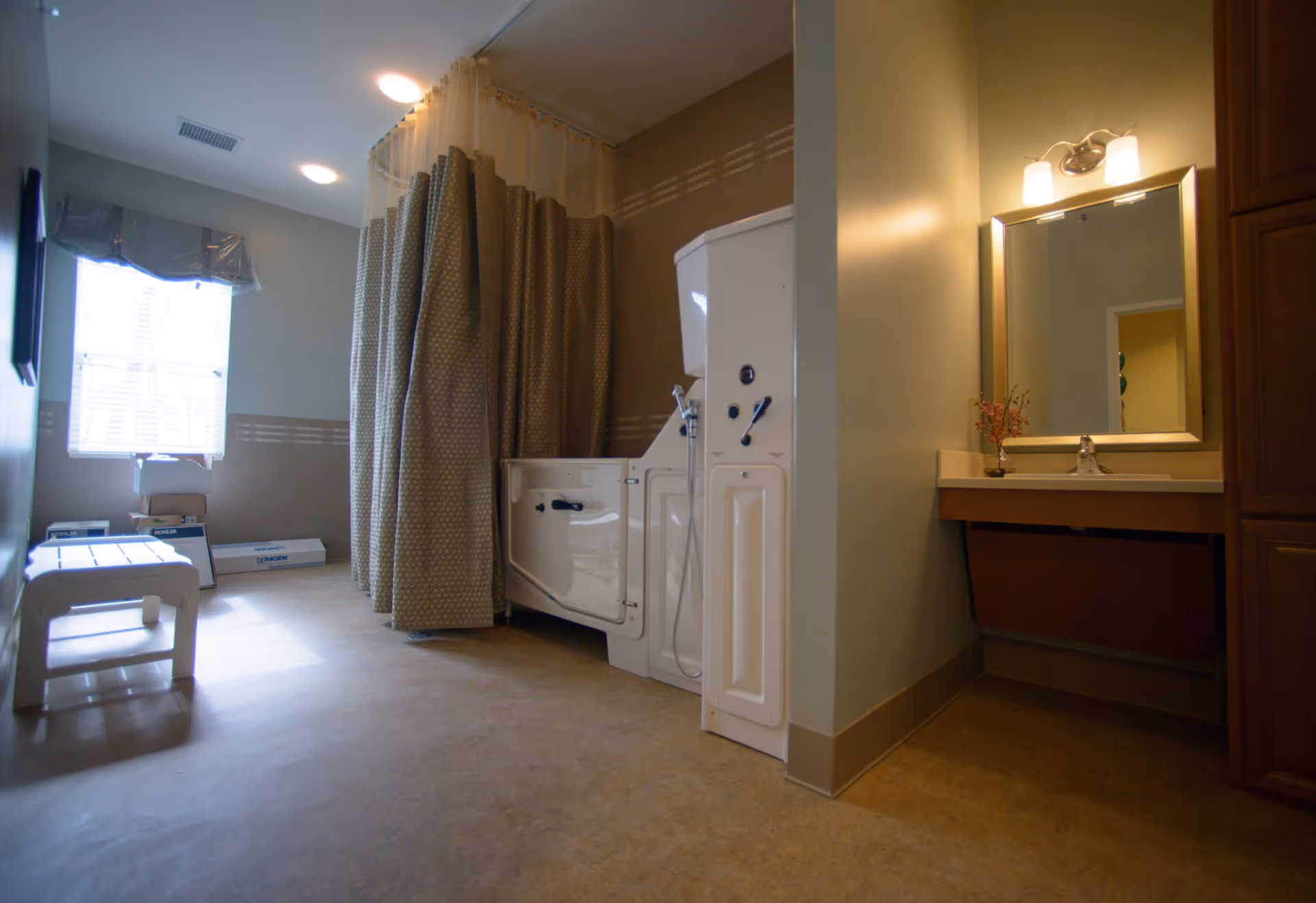 A spacious bathroom with a walk-in bathtub enclosed by a shower curtain, a vanity with a mirror and light fixture above it, a window with blinds, and a white bench near the window. The room has beige walls and flooring.