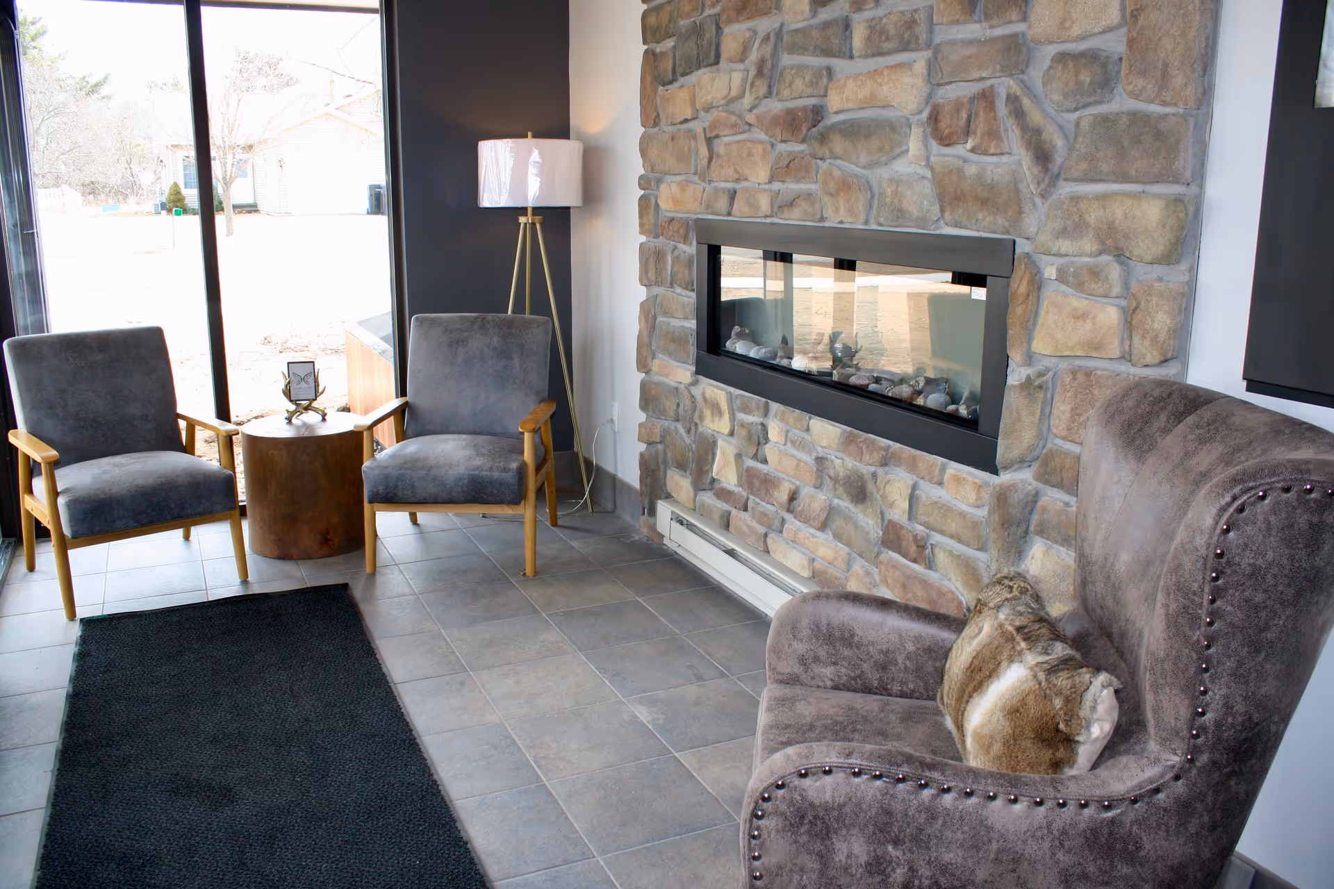 A cozy sitting area with a stone fireplace, two gray cushioned chairs with wooden arms around a small round wooden table, a tall floor lamp, and a plush armchair with a fur pillow. Large windows provide natural light and a view of the outdoors.