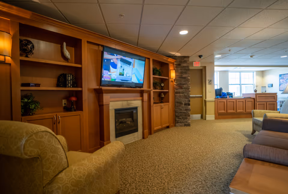 Common area with built-in wooden cabinets and fireplace, a wall-mounted TV, upholstered chairs, and a reception desk in the background.