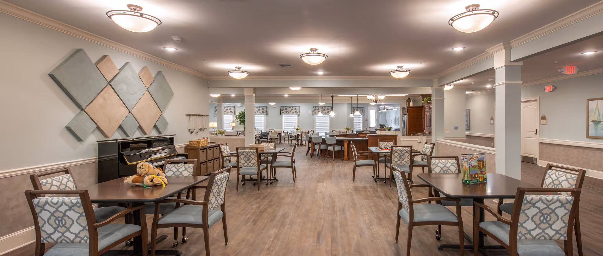 Spacious communal dining and activity room with multiple tables and chairs, a piano on the left, and a kitchenette/serving area in the back.