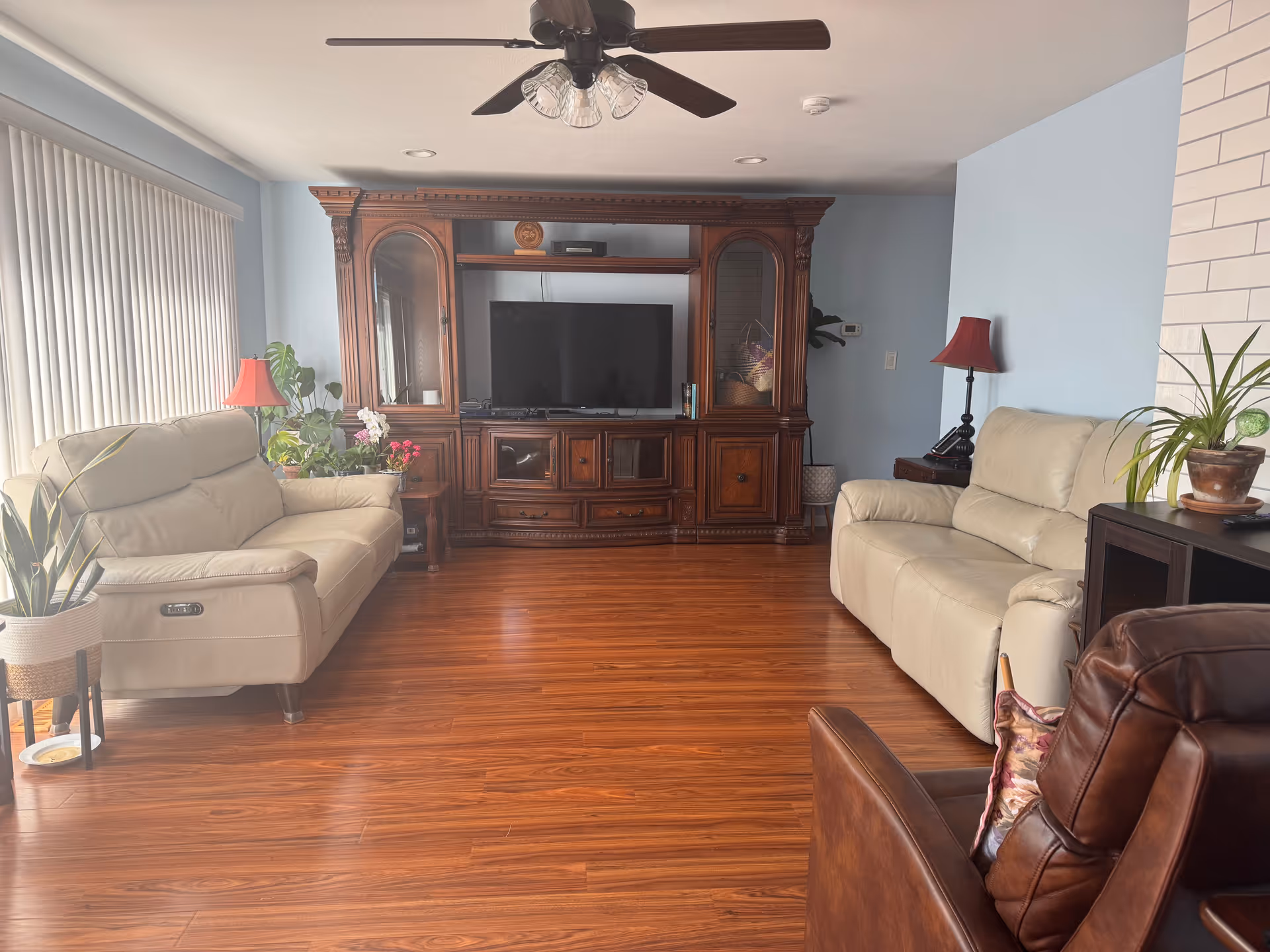 A cozy living room with wooden flooring, two beige leather sofas facing a large wooden entertainment center with a flat-screen TV. There is a brown leather armchair in the foreground, several potted plants, two red lamps, and a ceiling fan with lights above.