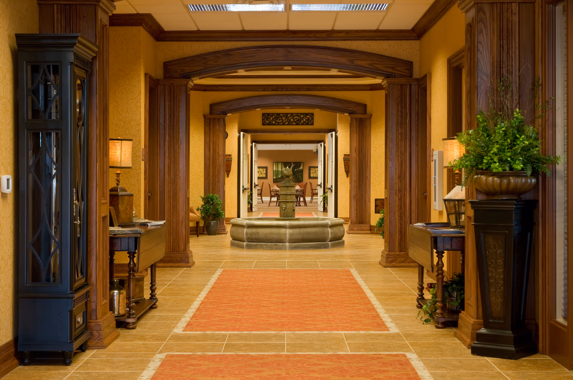 A warmly lit hallway with wooden archways and columns, featuring a central stone fountain and orange rugs on a tiled floor. The hallway is decorated with plants, lamps on side tables, and framed artwork in the background.
