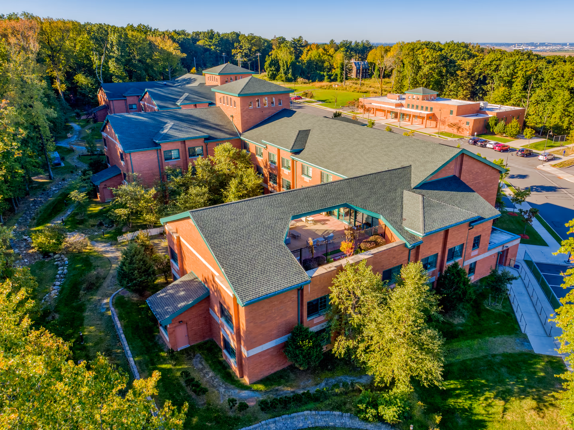 Aerial view of a large brick building complex surrounded by trees and greenery, with a parking lot visible on the right side and a clear blue sky above.