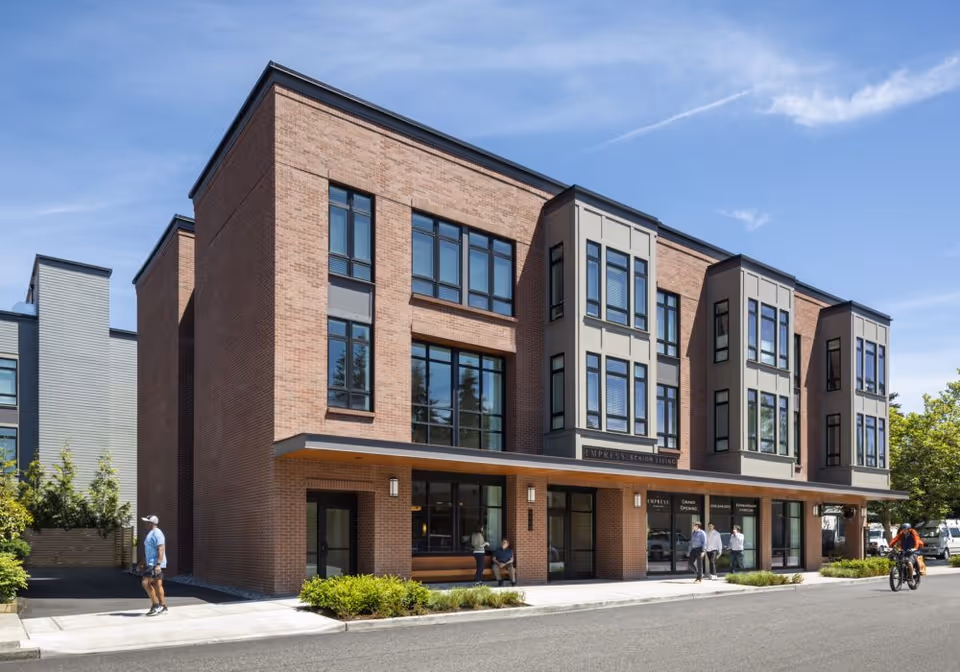 Exterior view of a modern three-story brick and gray panel building with large windows, identified as Empress Senior Living. People are walking and sitting near the entrance on a sunny day with a clear blue sky.