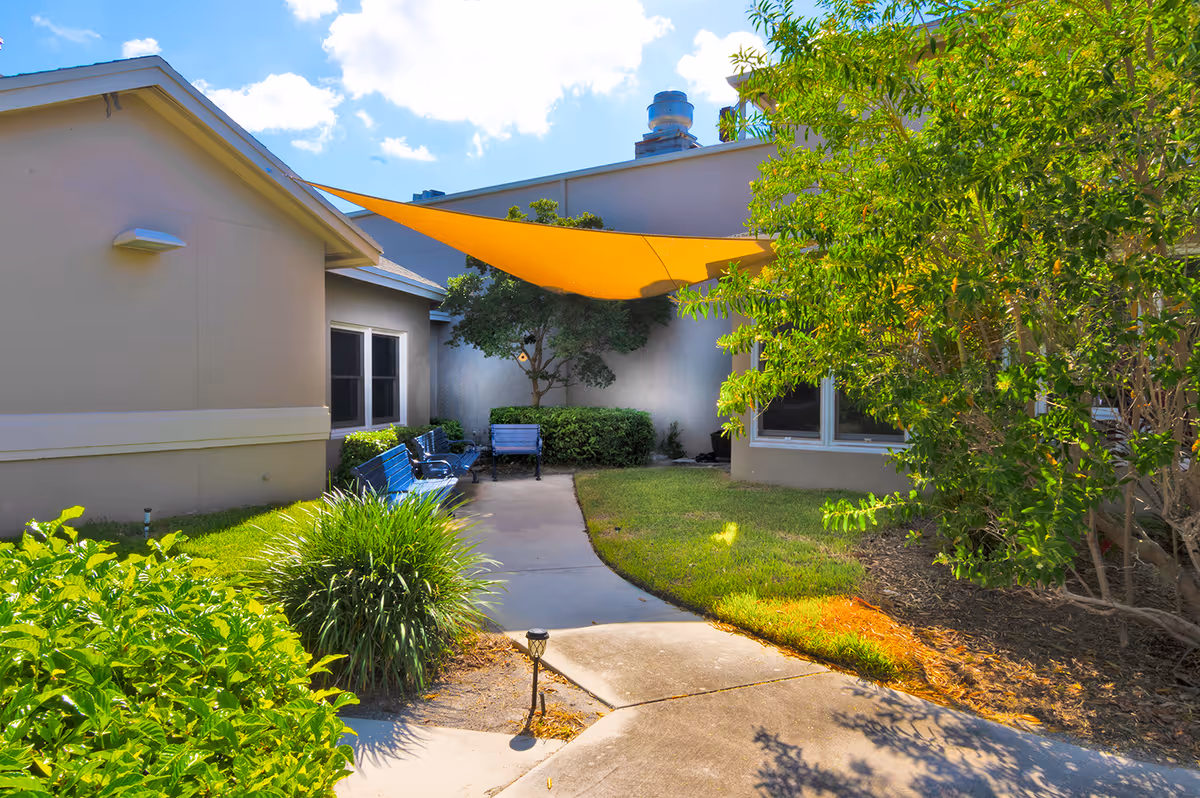 Outdoor courtyard area with a concrete pathway, green bushes, and trees. There are blue benches along the pathway and a large orange shade sail providing partial cover. The building walls are beige with windows visible, and the sky is blue with some clouds.