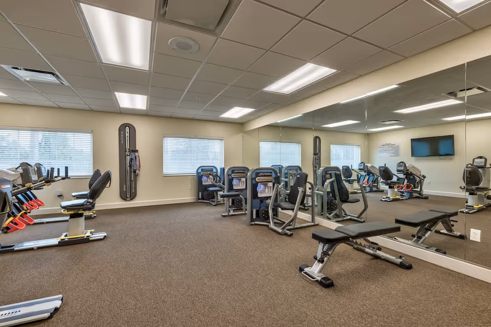 A fitness room with various exercise equipment including stationary bikes, weight machines, and a bench. The room has large mirrors on one wall, windows with blinds, and a wall-mounted TV.