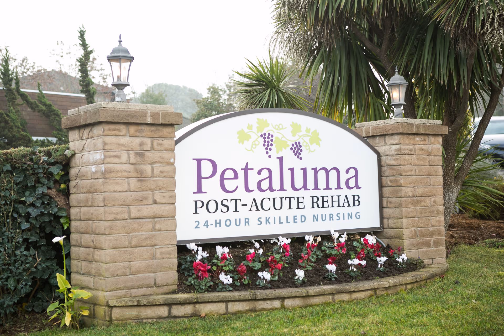 Outdoor sign for Petaluma Post-Acute Rehab with 24-hour skilled nursing, surrounded by flowers and greenery, with two brick pillars topped with lanterns on either side.