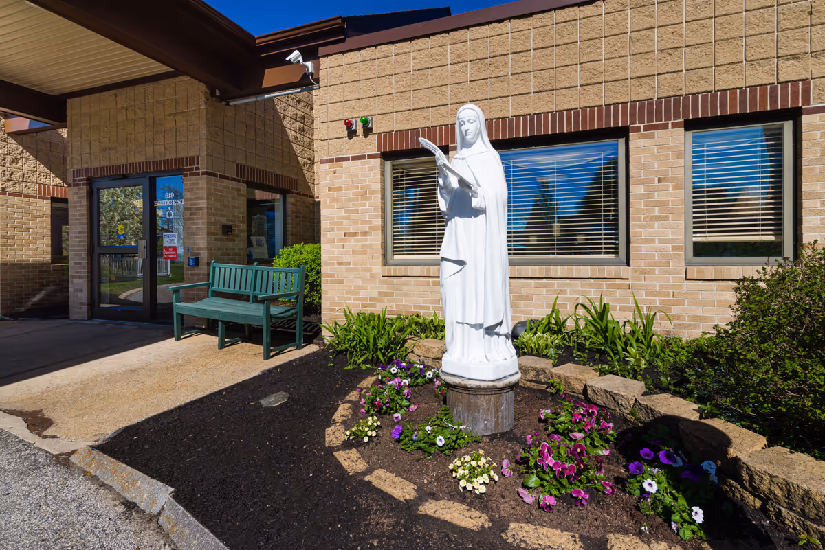 Outdoor view of the entrance to a building with a green bench near the door and a white statue of a woman reading a book surrounded by flowers and plants in a garden bed.