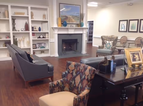 A cozy living room area with hardwood floors, featuring a gray sofa, patterned armchairs, a dark wooden table with framed photos, a white fireplace with decorative vases and a landscape painting above it, and built-in white shelves with various decorative items.