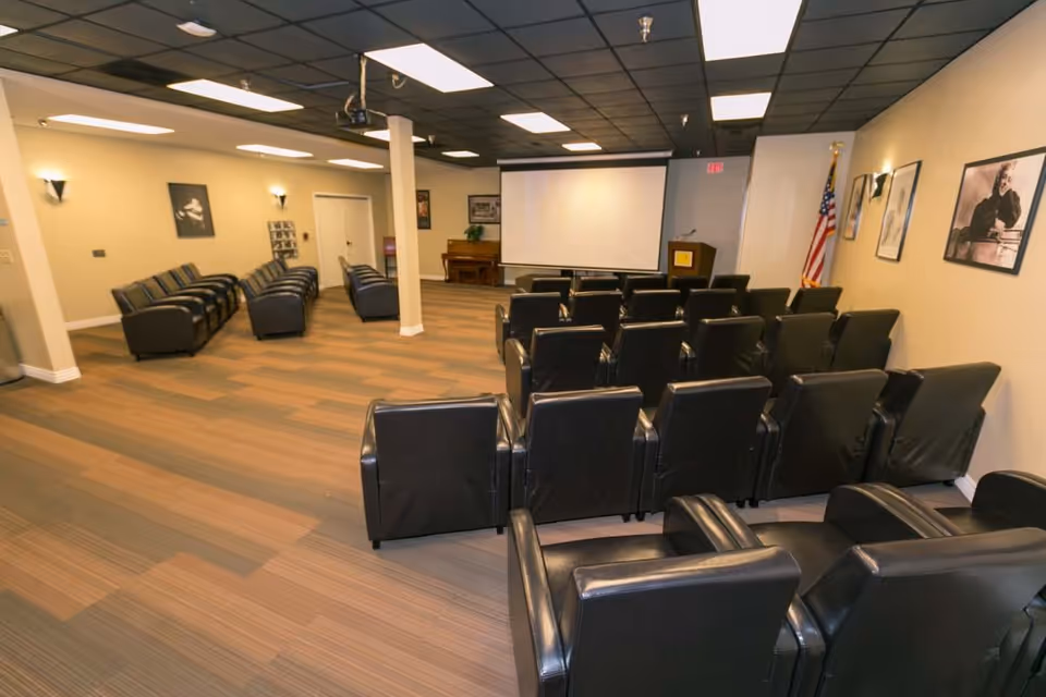 A large room set up as a small theater or presentation area with multiple rows of black leather chairs facing a white projection screen. There is a podium with a microphone and an American flag in the corner. The walls are decorated with framed black and white photographs, and there is a piano against the back wall.