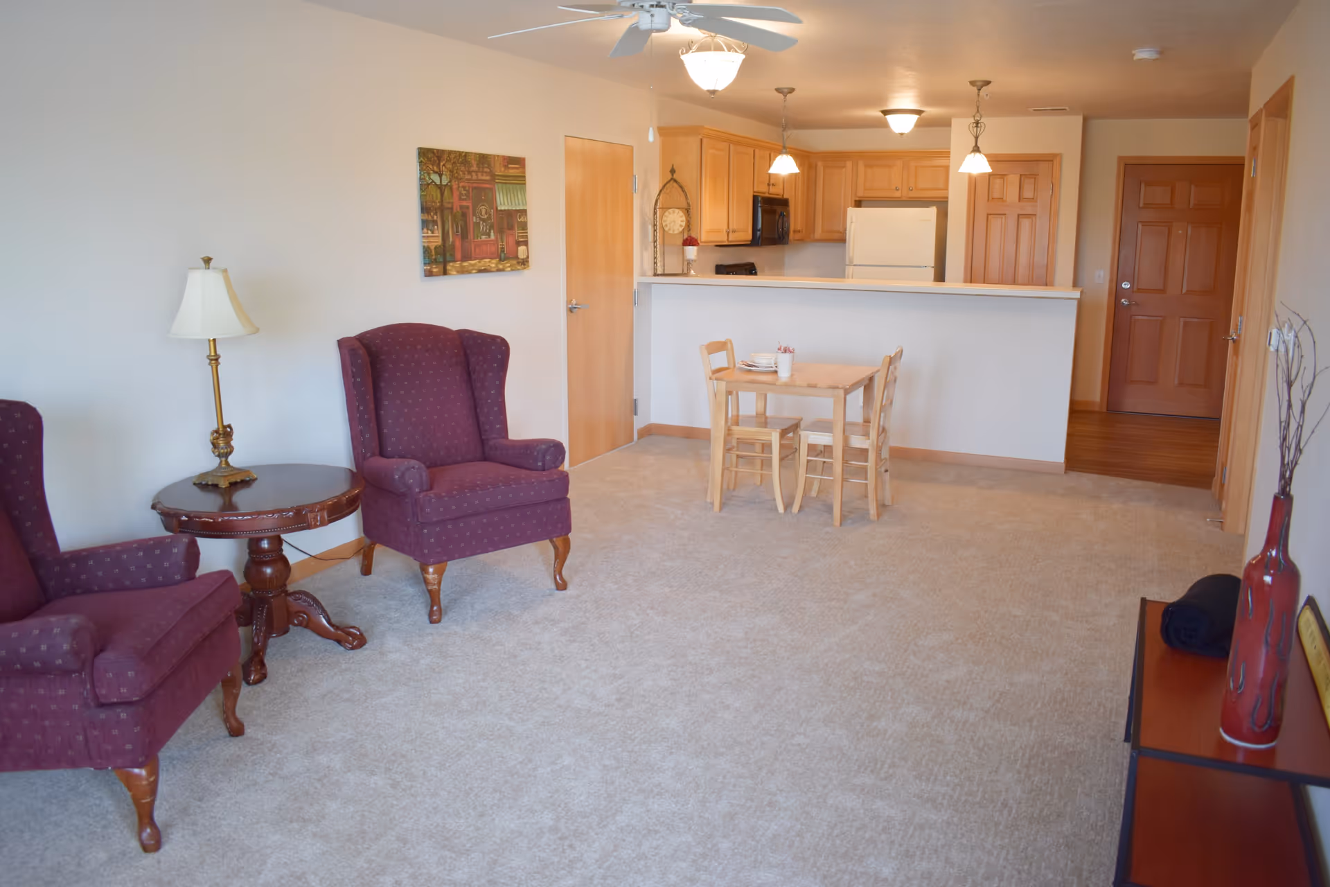 A cozy living area with two purple upholstered armchairs and a round wooden side table with a lamp. In the background, there is a small wooden dining table with two chairs, and a kitchen with wooden cabinets, a refrigerator, and pendant lights hanging from the ceiling. The room has beige carpet and light-colored walls with a painting and a ceiling fan.