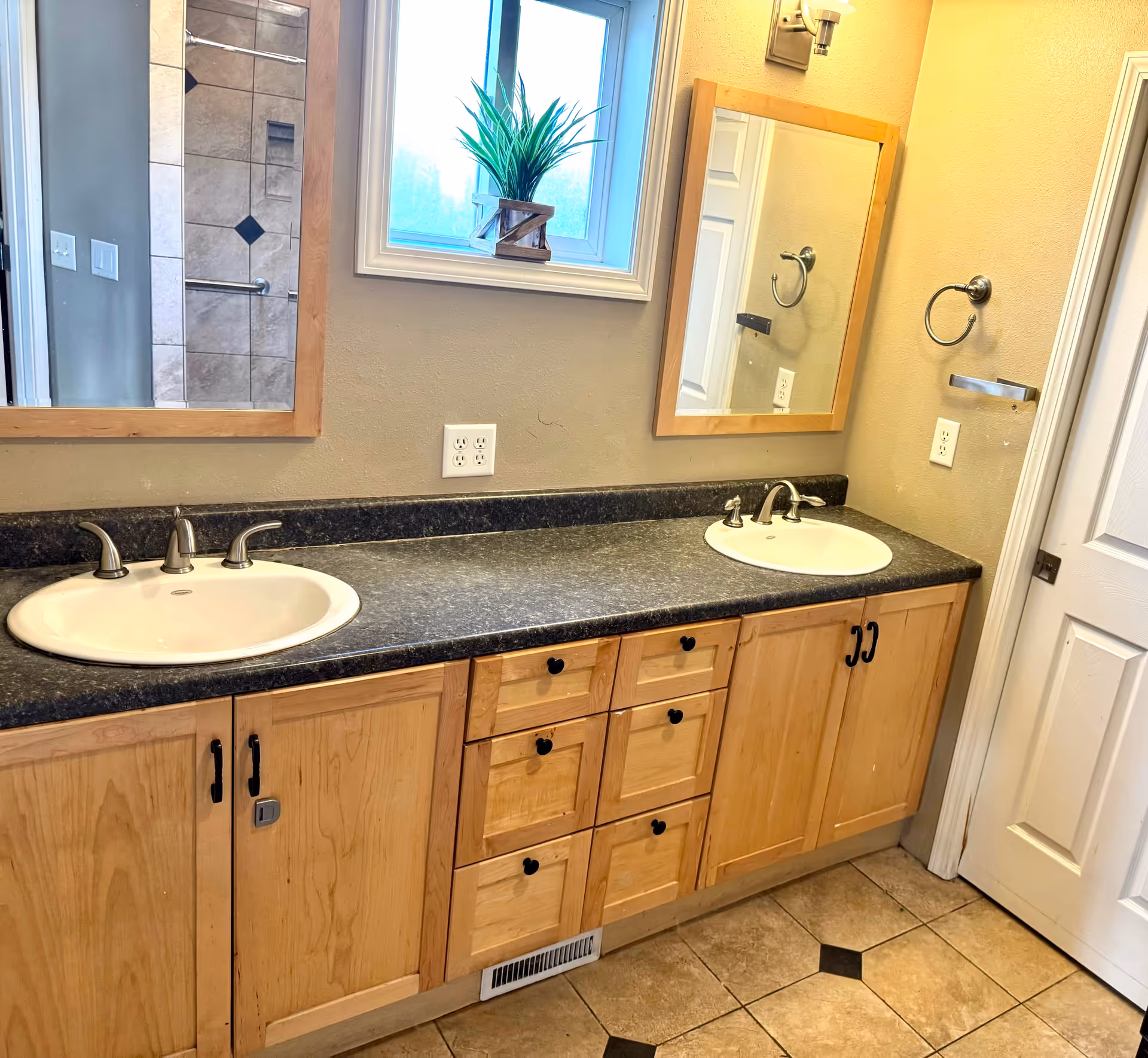 A bathroom interior featuring a double sink vanity with light wood cabinets and black countertops. Above each sink is a rectangular mirror with a wooden frame. A small window with a potted plant is situated between the mirrors. The floor is tiled with a beige and black diamond pattern. A towel ring and a soap holder are mounted on the wall near the right sink.