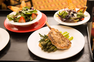 A plated meal featuring a grilled pork chop garnished with rosemary, served on a bed of asparagus, accompanied by two bowls of fresh salad with mixed greens and sliced strawberries.