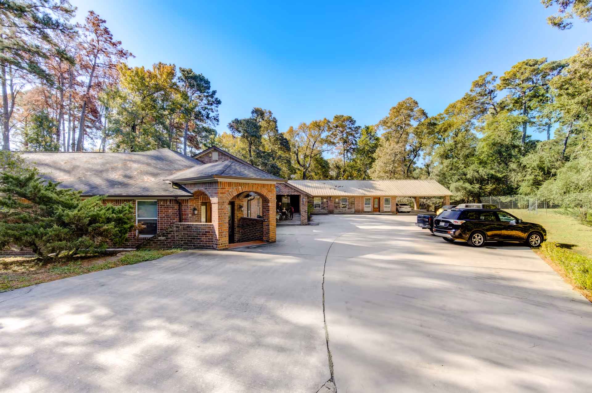 Exterior view of Forest Cottage Senior Care in Willis, Texas, showing a single-story brick building with a covered entrance and a spacious concrete driveway with parked cars. The building is surrounded by trees and greenery under a clear blue sky.